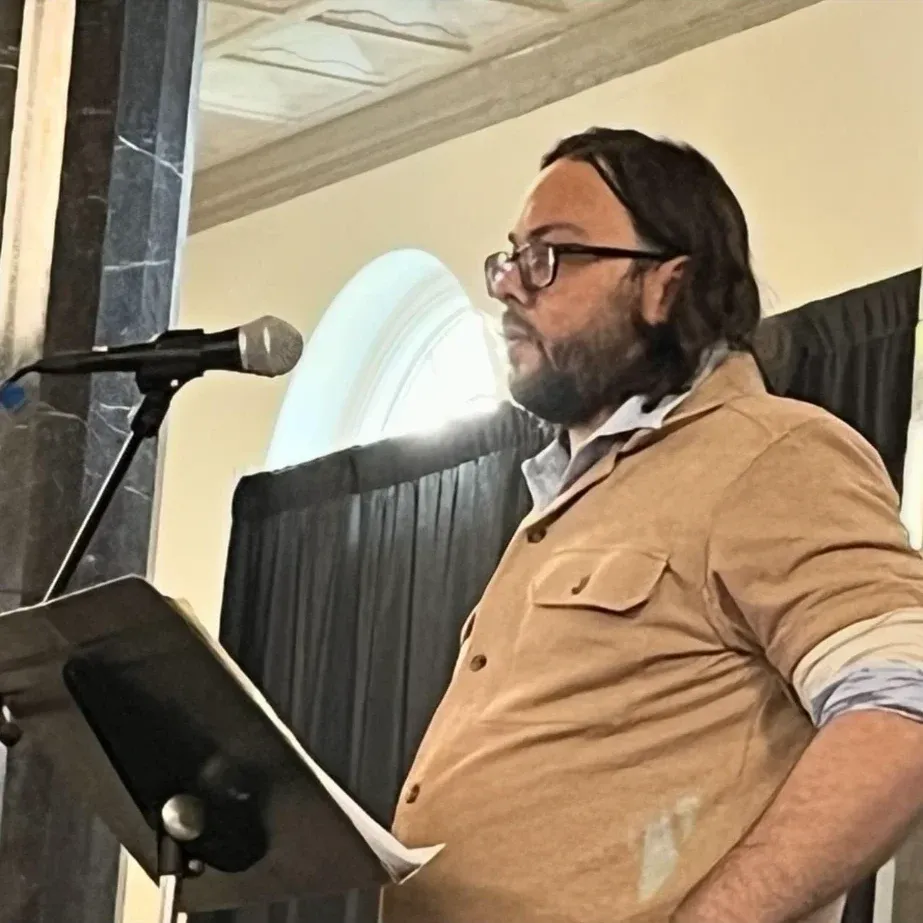 Eric Shay Howard reading from a lectern at a microphone in a tan shirt and glasses.