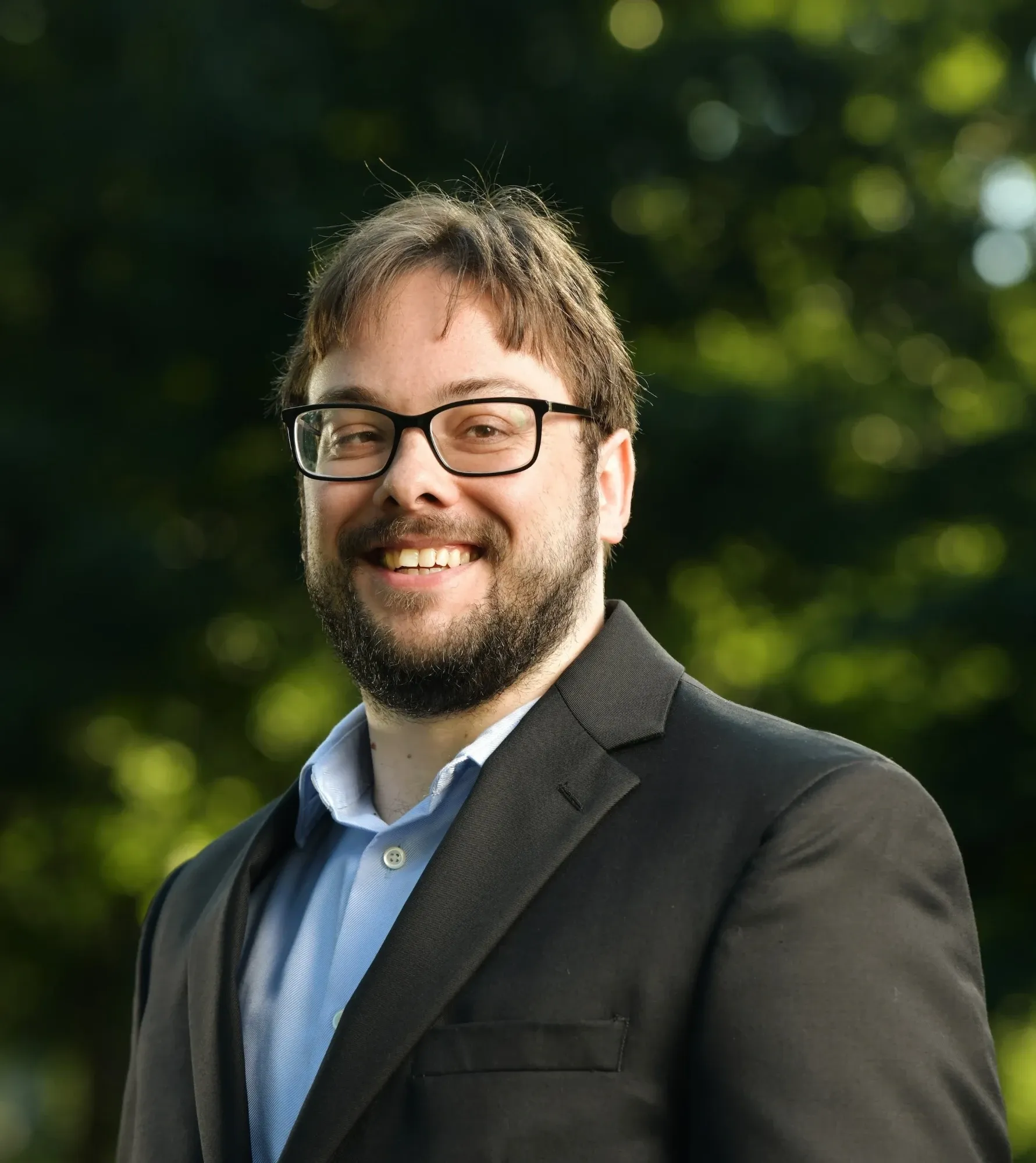 Eric Shay Howard smiling outdoors in a dark blazer and light blue shirt, trees glowing behind him.